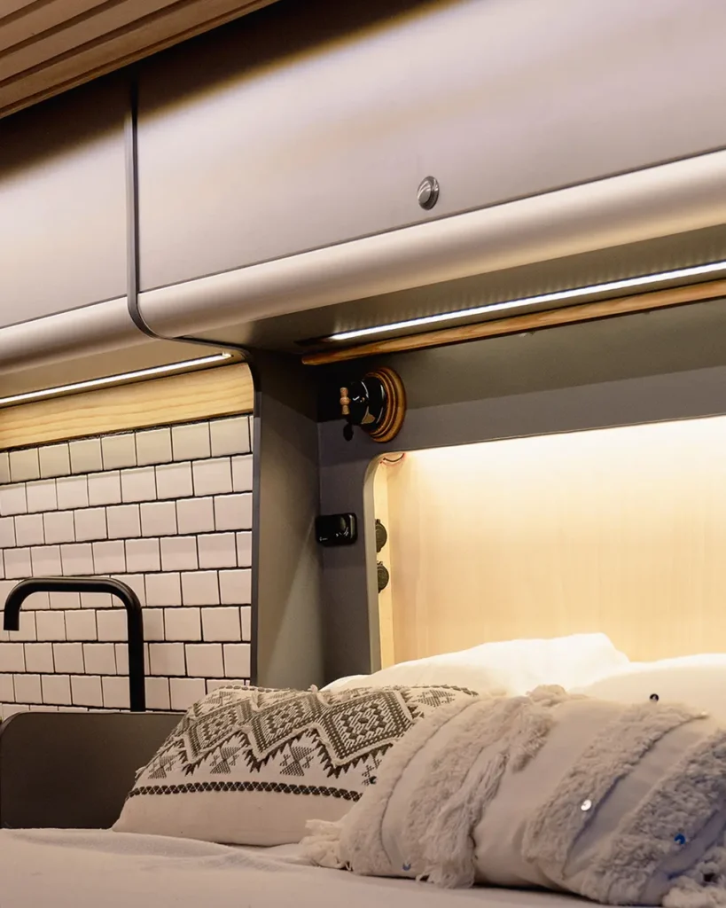 Área de descanso en la Fiat Ducato convertida, con una cama grande equipada con ropa de cama acogedora y un sistema de iluminación cálido integrado en la estructura superior.