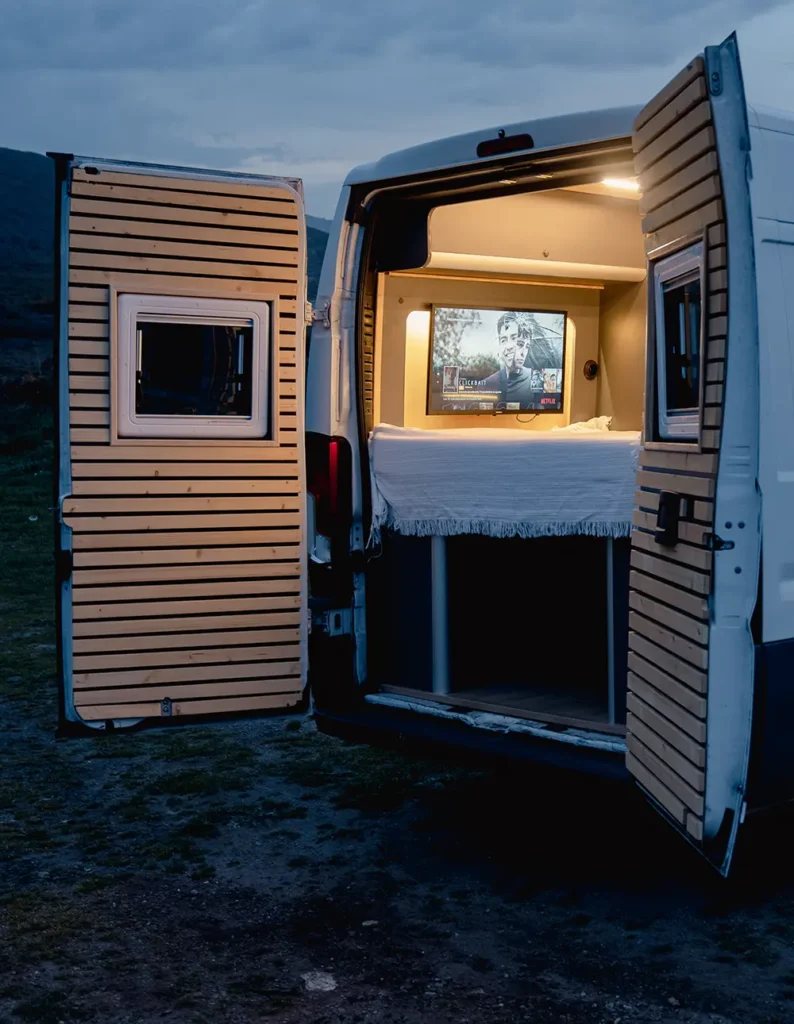 Vista nocturna de la parte trasera de una Fiat Ducato abierta, iluminada por luces interiores que resaltan un área de descanso con pantalla de televisión mostrando una película, con el paisaje natural al fondo.