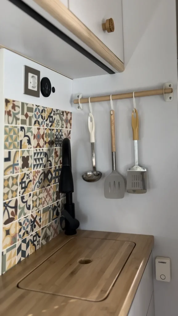 Vista interior de una cocina moderna en una furgoneta Fiat Ducato, equipada con armarios superiores de color blanco, encimera de madera, y diversos utensilios de cocina colgando en una pared decorativa de azulejos multicolor.