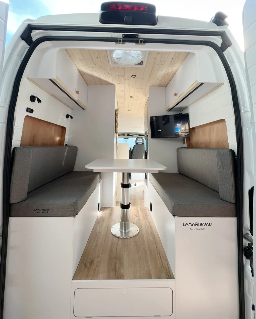 Detalle de la esquina de una mesa de madera clara y los bancos tapizados en gris dentro de una furgoneta Fiat Ducato, enfocando los acabados y la calidad del mobiliario.