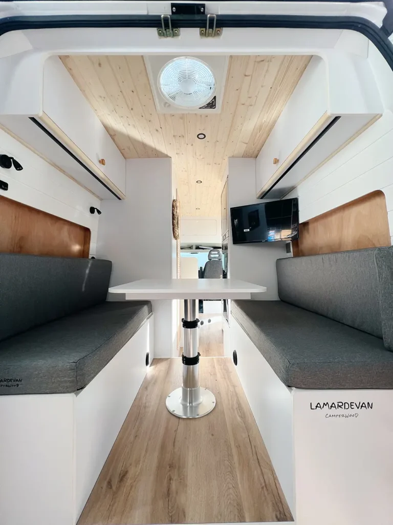 Vista trasera del interior de una furgoneta Fiat Ducato, mostrando la disposición de los asientos y la mesa del comedor con la cabina y el acceso al vehículo al fondo.