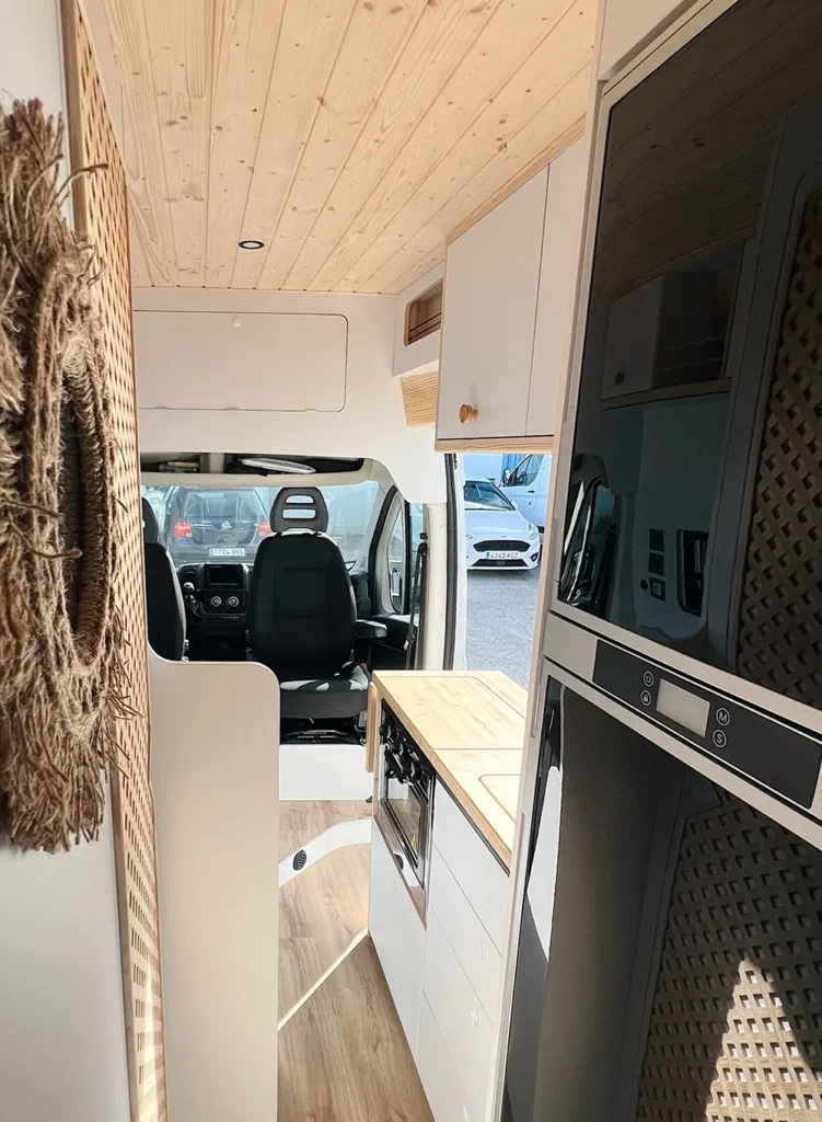 Vista superior del área de comedor en una furgoneta Fiat Ducato, con una mesa blanca y bancos laterales grises, iluminada por luz natural del techo.