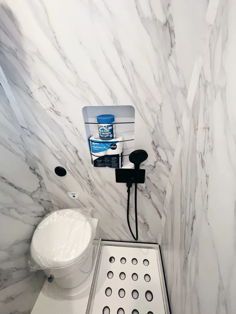 Baño compacto en una furgoneta Fiat Ducato con paredes de mármol falso, mostrando un inodoro moderno y una ducha con accesorios contemporáneos.