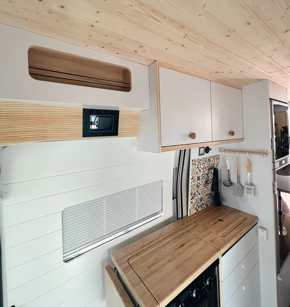 Detalle de una cocina en una furgoneta Fiat Ducato, mostrando una encimera de madera con un fregadero incorporado y una pared de azulejos con utensilios de cocina colgados