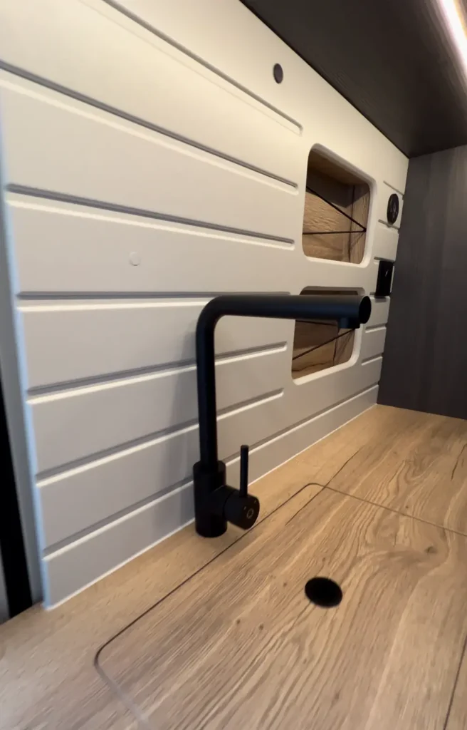 Espacio de cocina en una furgoneta Fiat Ducato, equipado con encimera de madera y grifo moderno, iluminado por luces LED, reflejando un ambiente cálido y acogedor.
