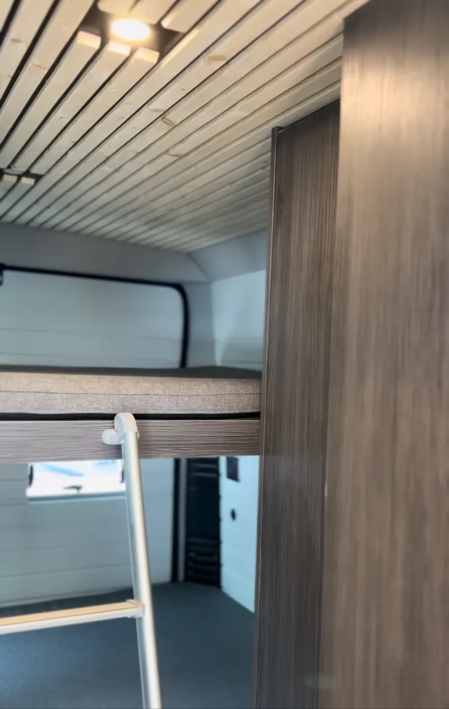 Vista lateral de una cama elevada y la zona de entrada en una furgoneta Fiat Ducato, con detalles del diseño interior optimizado para maximizar el espacio y la comodidad.