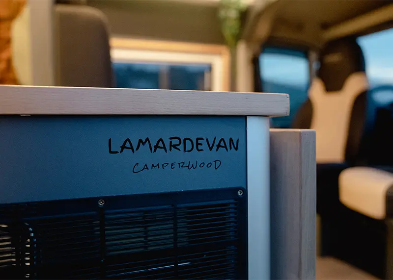 Interior de una furgoneta con acabados en madera, mostrando un mueble azul oscuro con el logo de 'LAMARDEVAN CAMPERWOOD'.