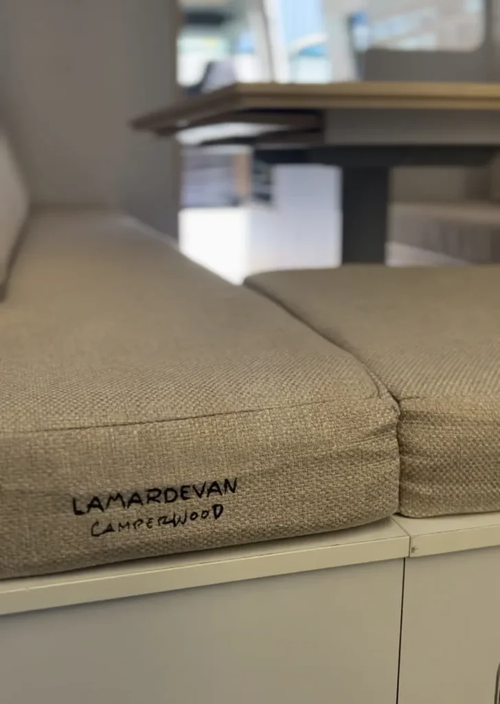 Imagen del interior de una furgoneta VW Crafter convertida en camper, mostrando un sofá-cama de tela gris con la etiqueta "LAMARDEVAN Camperwood" visible.
