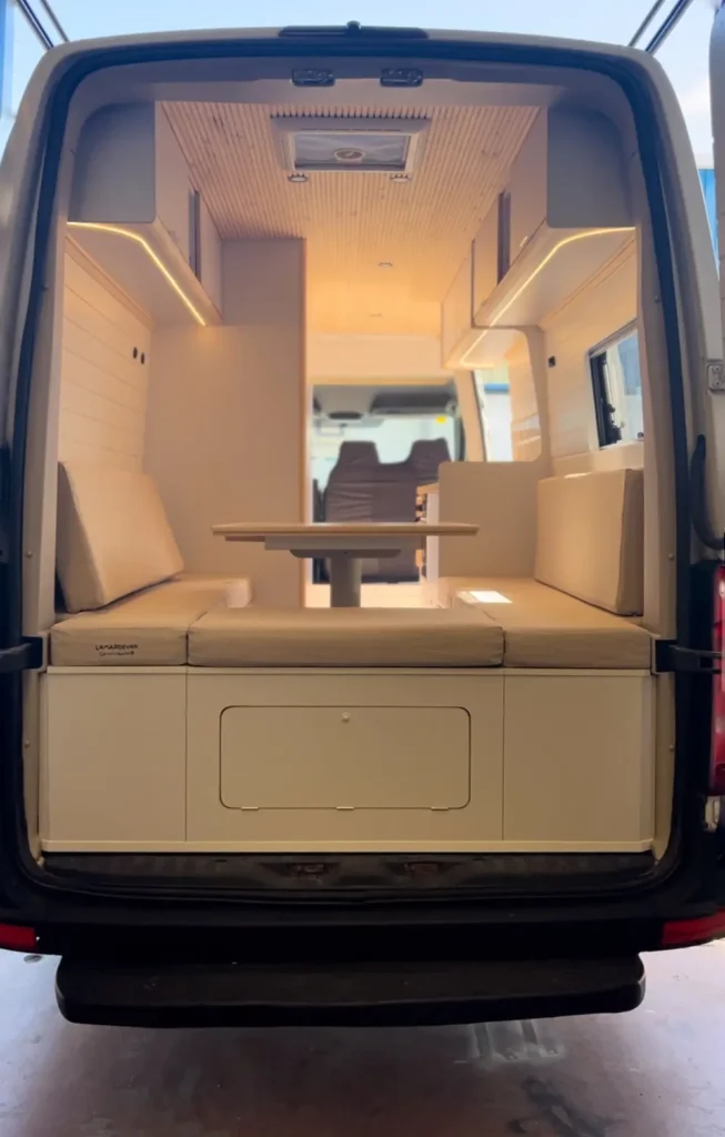 Vista trasera de una furgoneta VW Crafter convertida en camper con las puertas traseras abiertas, revelando un interior bien iluminado con asientos y una mesa plegable.