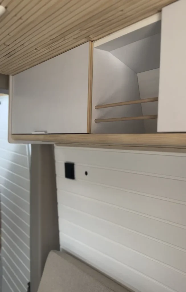Detalle del almacenamiento superior en una furgoneta VW Crafter convertida en camper, mostrando un armario de puerta plana de color blanco con tiradores de madera.