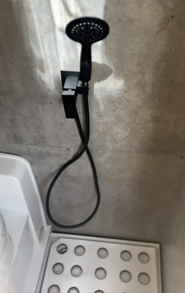 Imagen del área de ducha en una furgoneta VW Crafter convertida en camper. Se muestra una ducha moderna con cabezal negro y paredes de efecto concreto, proporcionando un aspecto industrial al espacio.
