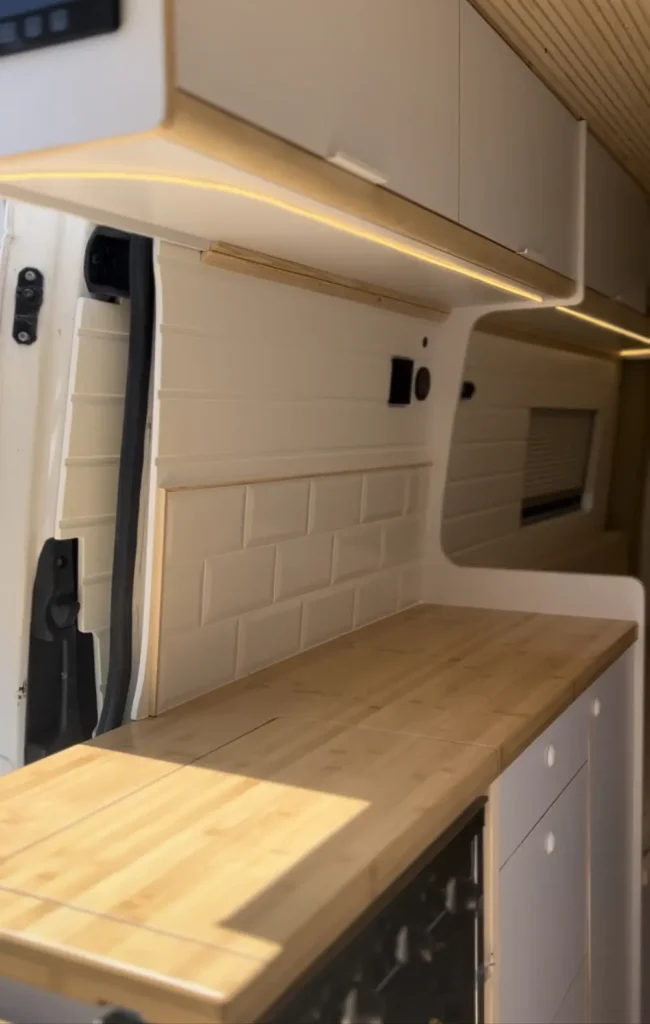 Imagen de una furgoneta Fiat Ducato L3H3 mostrando una cocina compacta y moderna. Se destaca la encimera de madera y los armarios blancos, junto a una ventana con persiana enrollable.