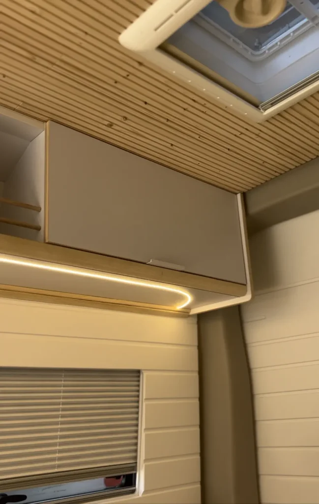 Vista del área de almacenamiento y techo en una furgoneta VW Crafter convertida en camper. La imagen muestra armarios superiores, una escotilla en el techo y una iluminación suave, creando un ambiente acogedor dentro de la camper.